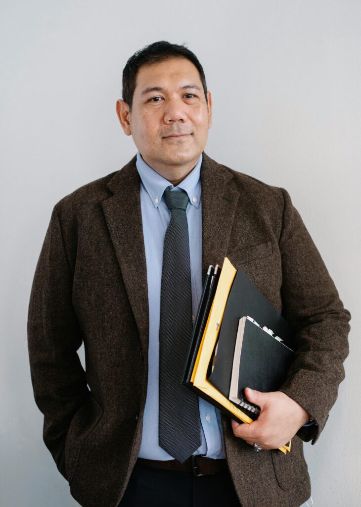 Positive Asian male office worker in formal suit with paper folders in hand standing with hand in pocket against white wall